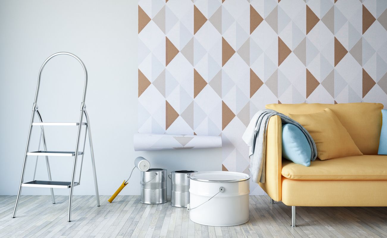 Ein Raum mit einer gelben Couch, einer Leiter, Farbrollen und einem Eimer auf einem Holzboden. Die Wand hat ein geometrisches Muster in Weiß und Braun.