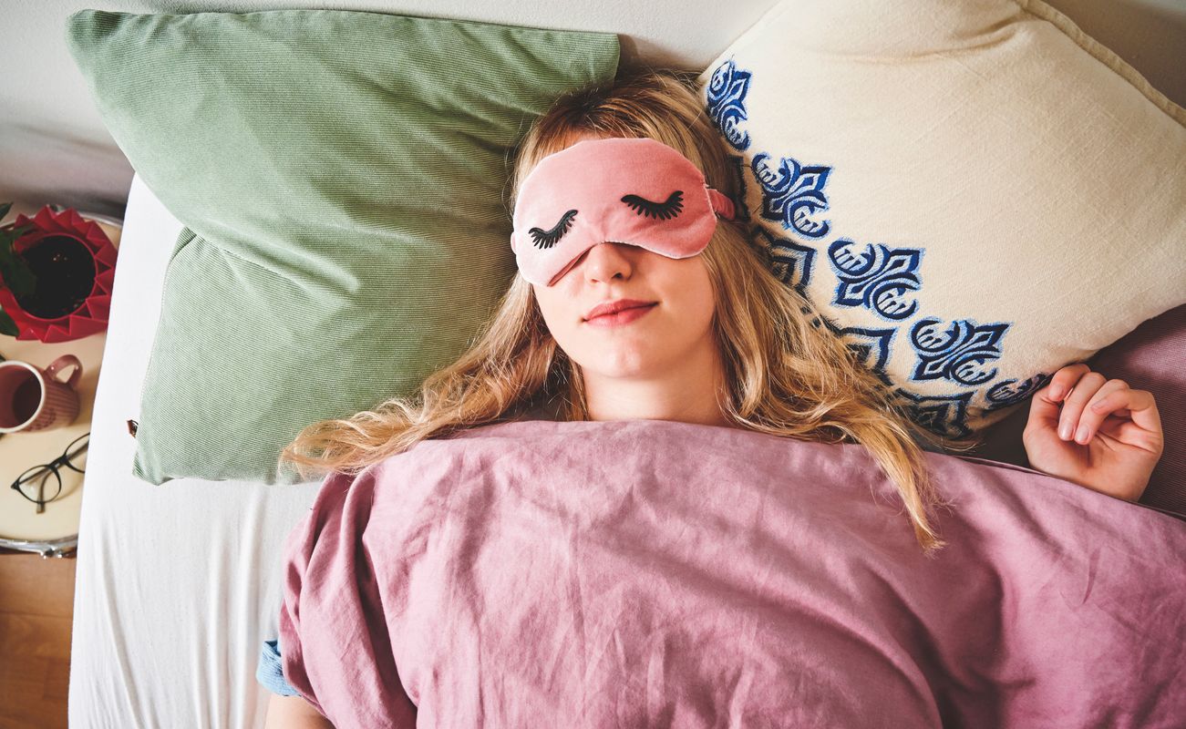 Eine schlafende Person mit einer Schlafmaske und langen, blonden Haaren, umgeben von Kissen und einer Decke.